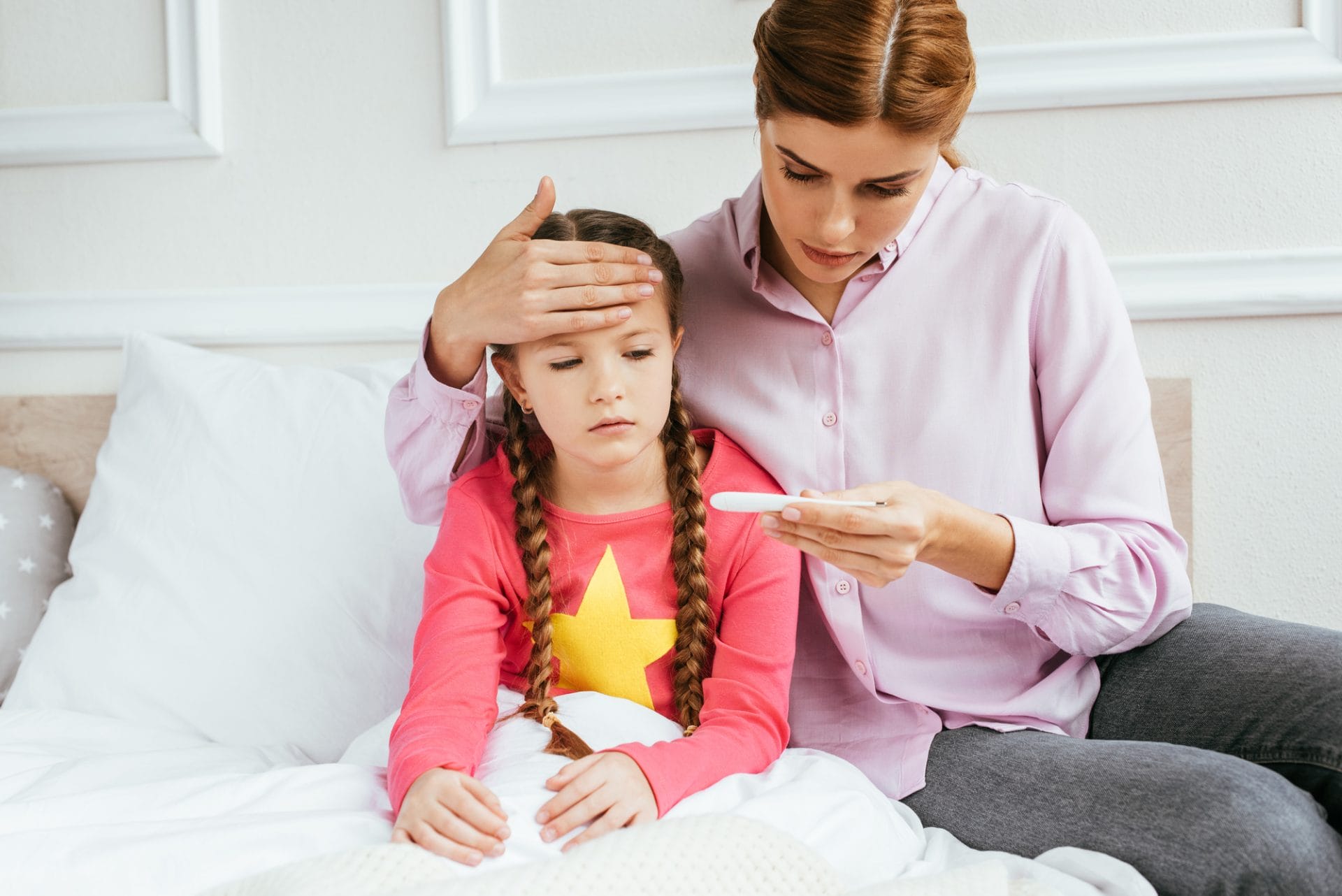 A mother checking her daughter's temperature with a thermometer while the child rests in bed.
