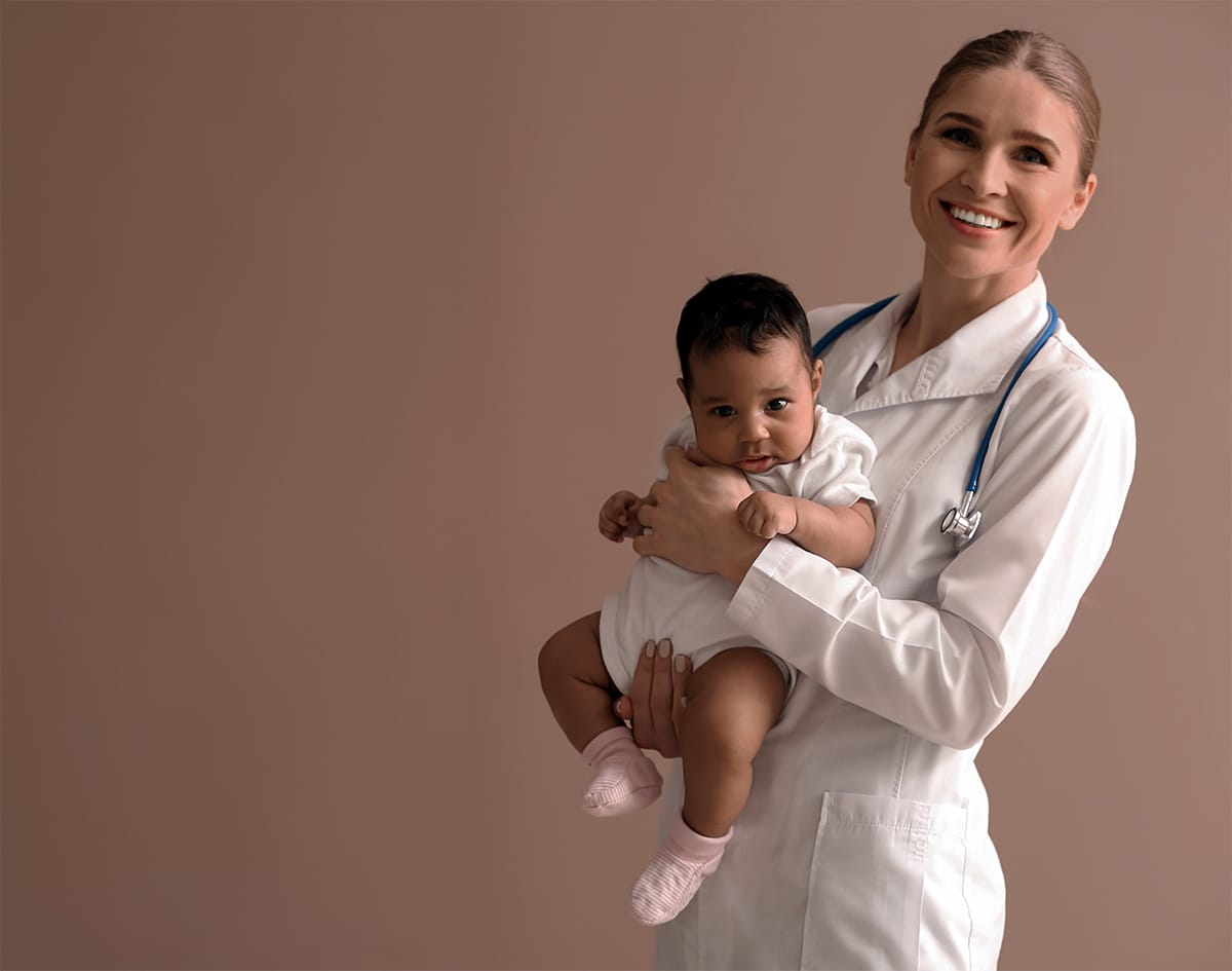 A smiling healthcare professional holding a baby with care and confidence against a neutral background.