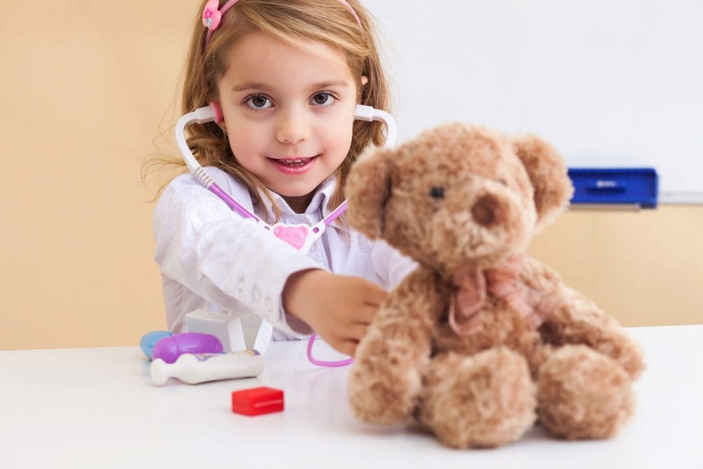 A young girl plays pretend doctor with a stethoscope and a teddy bear.