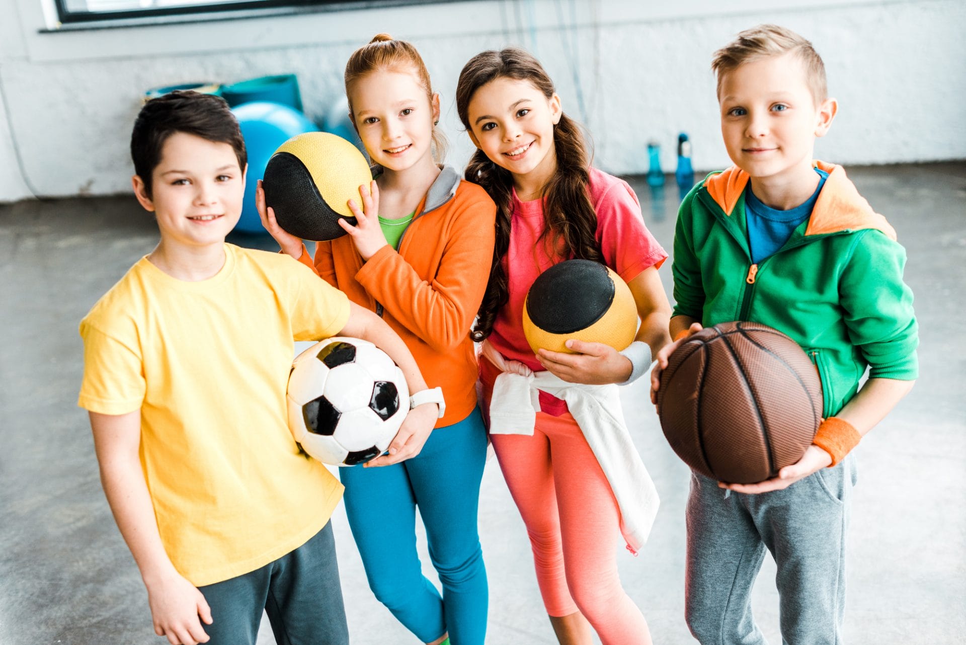 Four children holding various sports balls and smiling at the camera.
