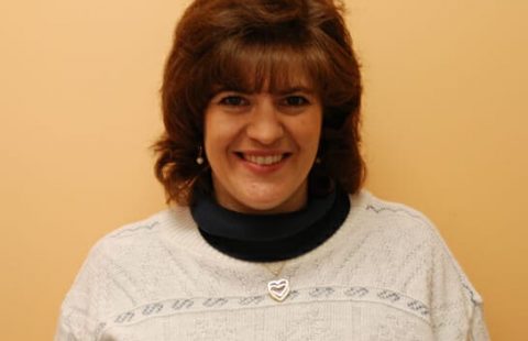 Woman with a smile wearing a white sweater with a navy collar and a heart-shaped pendant.