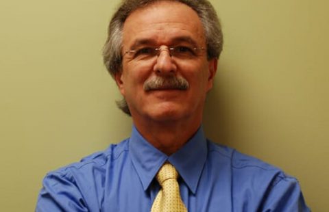 A man with a mustache wearing a blue shirt and a yellow tie, smiling in front of a neutral-colored wall.