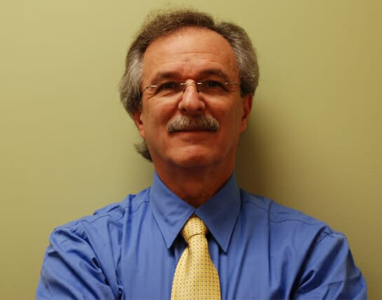 A man with a mustache wearing a blue shirt and a yellow tie, smiling in front of a neutral-colored wall.