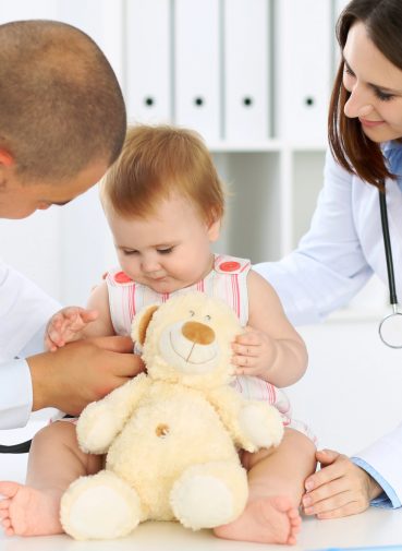 Two pediatricians interacting with a baby patient holding a stuffed toy.