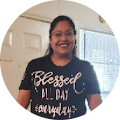 A smiling woman wearing glasses and a black t-shirt with the phrase "blessed all day everyday" standing indoors.
