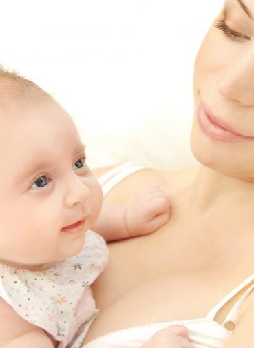 A woman gazes lovingly at the baby in her arms, who looks upwards with a curious expression. They are both bathed in soft, natural light.
