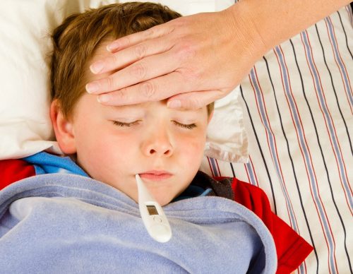 A child with a thermometer in his mouth appears to be sick while an adult hand is touching his forehead, likely checking for fever.