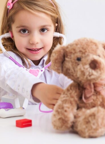 A young girl plays pretend doctor with a stethoscope and a teddy bear.