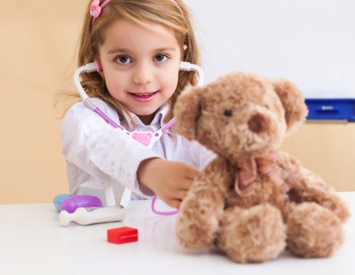 A young girl plays pretend doctor with a stethoscope and a teddy bear.
