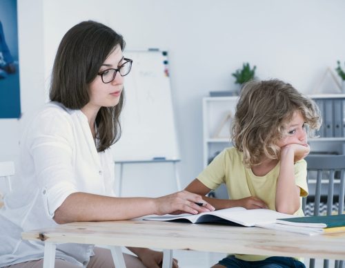 A woman assists a young child with homework at a white table in a room with shelves and educational posters.